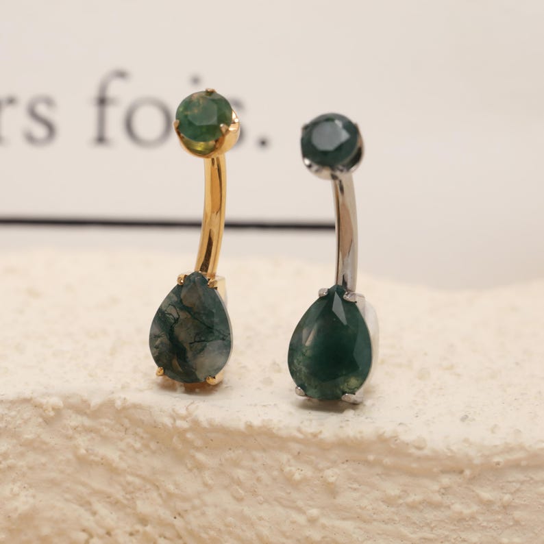May include: Two belly button rings. One has a gold-colored curved bar with a green gemstone at the top and a teardrop-shaped green gemstone at the bottom. The other has a silver-colored curved bar with a green gemstone at the top and a teardrop-shaped green gemstone at the bottom.