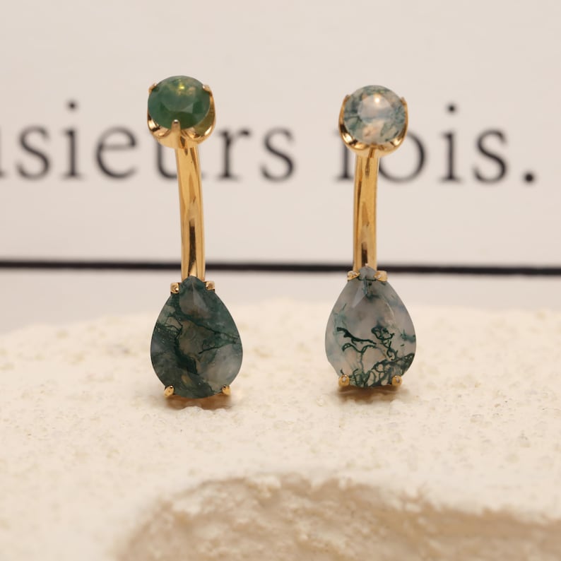 May include: A pair of gold-colored earrings with a teardrop-shaped, moss agate stone. Each earring has a round, green stone at the top. The earrings are displayed on a white surface.