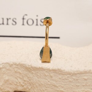 May include: A gold-colored earring with a teardrop-shaped green stone. The earring has a post with a small green stone at the top. The earring is set on a textured, off-white surface.