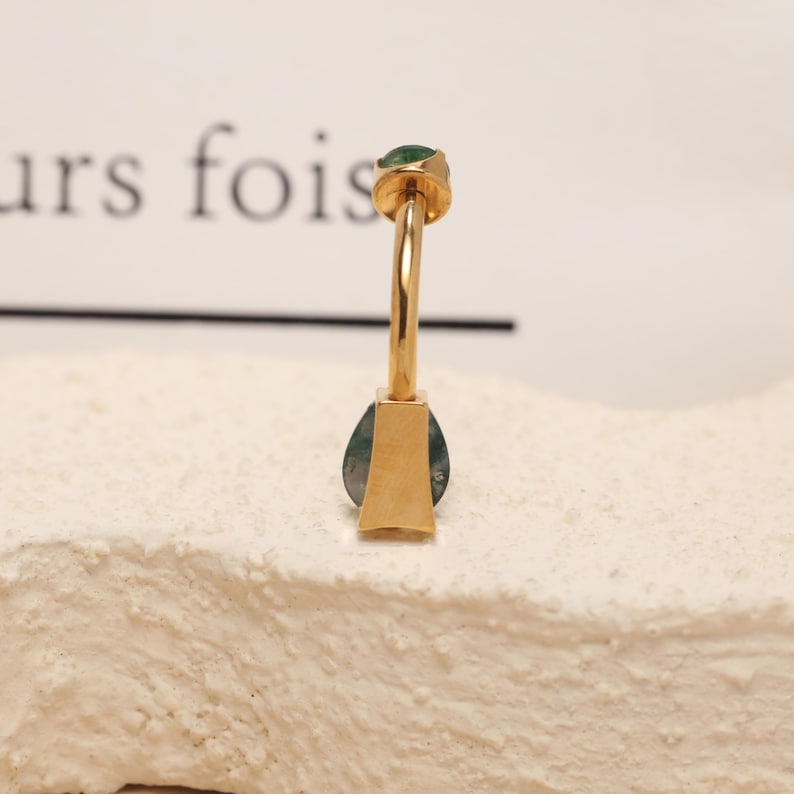 May include: A gold-colored earring with a teardrop-shaped green stone. The earring has a post with a small green stone at the top. The earring is set on a textured, off-white surface.