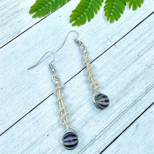 May include: Handmade silver wire earrings with a spiral design and a striped, dark purple and gray bead at the bottom. The earrings are displayed on a white wooden surface with green leaves in the background. The text "Brigitte Barnett" is at the bottom.