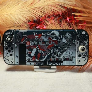 May include: A black and grey gaming console skin featuring Persona 5 character graphics, white star patterns, and Chinese text. The device includes two controller attachments with paw print thumb grips and a central screen display area.