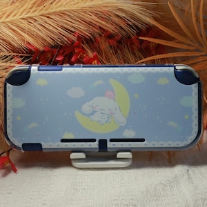 Op de afbeelding: Een lichtblauwe beschermhoes voor een console met een wit stripfiguur dat op een gele halve maan slaapt. Het ontwerp bevat kleine witte wolken, sterren en een delicaat kantachtig randpatroon langs de randen van de rechthoekige behuizing.