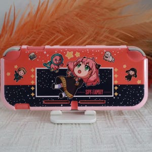 Könnte beinhalten: Eine rosa Schutzhülle für eine tragbare Spielkonsole mit Spy Family-Motiv. Das Design zeigt eine zentrale Illustration einer Figur mit rosa Haaren, kleinere Figuren-Icons, Sterne und den Schriftzug Spy Family vor einem dunkelblauen Sternenhimmel.