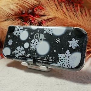 Könnte beinhalten: Eine schwarze Schutzhülle für eine tragbare Spielkonsole mit weißem Schneeflockenmuster und kreisförmigen Bokeh-Punkten. Die transparente Schutzhülle zeigt das Nintendo Switch-Logo und regulatorische Texte auf der Rückseite, auf einem Ständer.