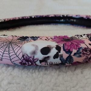 May include: Pink steering wheel cover with a floral and skull design. The design features pink flowers, a spiderweb, and white skulls.
