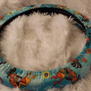 May include: A turquoise blue steering wheel cover with a colorful Native American design. The cover features stylized figures and geometric patterns in shades of orange, yellow, and brown.