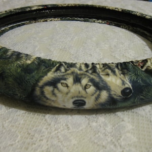 May include: A steering wheel cover with a wolf print design. The cover is made of a soft, plush fabric and features a green and brown color scheme. The wolf print is realistic and detailed, with the wolves' eyes and noses clearly visible.
