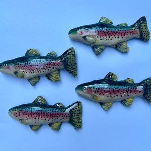May include: Four ceramic rainbow trout ornaments. Each fish has a green back, a red and pink stripe, and yellow fins. The trout are arranged on a light blue background. The fish are detailed with black spots.