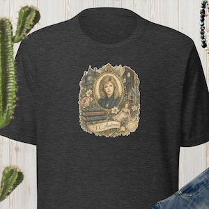 May include: A dark charcoal gray short-sleeve t-shirt featuring a central graphic illustration of a woman in an ornate gold frame, surrounded by books and flowers, with the text V.C. Andrews printed on a banner at the bottom of the design.