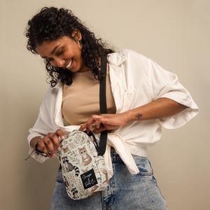 May include: A beige crossbody bag with a black strap. The bag has a woodland animal print with raccoons, foxes, and mushrooms. The bag has a black patch with the text "Freeper Wild Co."