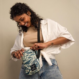 Könnte beinhalten: Eine kleine Umhängetasche mit weißem Hintergrund und einem blauen Wal-Druck. Die Tasche hat einen verstellbaren schwarzen Riemen und einen schwarzen Patch mit dem Text "Fin&Wild Co."