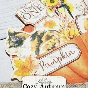 Puede incluir: Una tarjeta blanca con el texto "Cozy Autumn ephemera pocket" y unas tijeras con una pancarta que dice "Calico Collage Print-Cut-Create". La tarjeta est&aacute; decorada con un borde de encaje y una ilustraci&oacute;n de un gato marr&oacute;n y blanco. El fondo es una superficie de madera con varias manualidades de papel de tem&aacute;tica oto&ntilde;al, incluyendo un boleto que dice "Admit One" y una etiqueta que dice "Pumpkin".