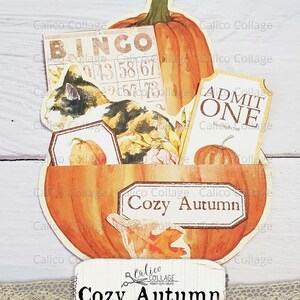 Puede incluir: Un bolsillo de ef&iacute;meras con forma de calabaza con una etiqueta "Cozy Autumn". El bolsillo est&aacute; lleno de ef&iacute;meras de papel con tem&aacute;tica oto&ntilde;al, incluyendo una tarjeta de bingo, un boleto "Admit One" y una ilustraci&oacute;n de un gato.