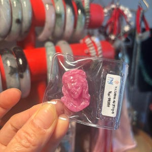Puede incluir: Un colgante de piedra preciosa rosa tallado en forma de rostro, sujeto en un paquete de plástico transparente. El colgante es de color rosa vibrante. Una pequeña etiqueta blanca con texto japonés es visible.