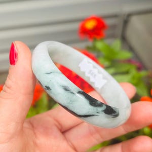 May include: A smooth, rounded bangle bracelet crafted from polished stone. The material is a pale, translucent off-white color featuring distinct, swirling veins of dark charcoal and deep forest green throughout the circular band structure.