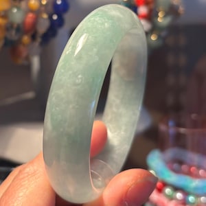 May include: A translucent jade bangle bracelet in shades of pale green. The smooth, polished surface reflects light, highlighting the natural variations in the stone. This elegant piece of jewelry is a classic accessory.