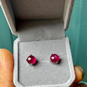 May include: A pair of round, cabochon-cut deep pink gemstone stud earrings set in silver-toned metal prongs. The earrings are displayed inside a light gray, velvet-lined jewelry box with a hinged lid, providing a neutral backdrop for the stones.