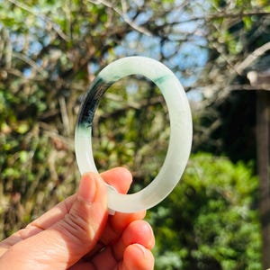 May include: A smooth, circular bangle bracelet crafted from translucent pale green jadeite. The bangle features a distinct, deep dark green to black mineral inclusion that creates a striking contrast against the lighter, milky green stone body.