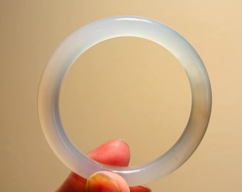 May include: A translucent, pale blue jade bangle bracelet. The smooth, circular bracelet is held by a hand against a neutral background. The jade has a subtle, natural variation in color.