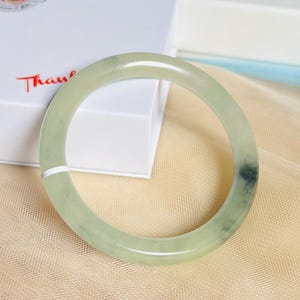 May include: A smooth, circular bangle bracelet crafted from pale green jade stone. The bangle features a polished, translucent surface with subtle natural variations in color, including a darker, mottled green inclusion on the right side.