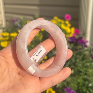 Könnte beinhalten: Ein glatter, durchscheinender hellrosa Rosenquarz-Armreif mit abgerundetem Profil. An der Innenseite ist ein kleines weißes rechteckiges Etikett befestigt, das den gedruckten Text trägt: NATURAL ROSE QUARTZ No: 100643.