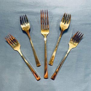 May include: Five gold forks arranged in a star shape on a blue surface.