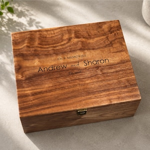 May include: A square wooden keepsake box with a natural brown wood grain finish. The lid is engraved with the text "OUR MEMORIES", "Andrew and Sharon", and "Est. 2026". A small gold-toned metal latch is centered on the front edge of the box.