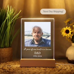 May include: A white LED light up photo frame with a picture of a man in a blue jacket. The frame has the text "A piece of our heart is in heaven DAD 1952-2024" on it.