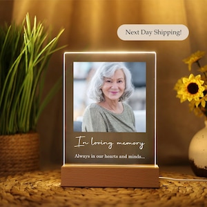 May include: A white acrylic photo frame with a wooden base. The frame has a photo of an older woman with a warm smile. The text "In loving memory" and "Always in our hearts and minds..." is printed on the frame.