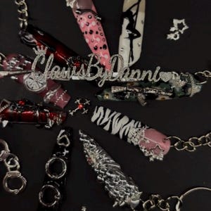 Op de afbeelding: Een assortiment versierde acrylnagels met verschillende ontwerpen, waaronder roze stippen, zebra strepen en een silhouet. Zilveren kettingen en bedels, waaronder handboeien en een kruis, zijn ook aanwezig. De tekst "Claws By Danni" wordt weergegeven.