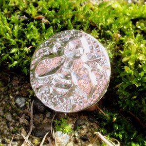 May include: A silver-colored metal pendant with a detailed bee design. The pendant is sitting on a bed of green moss.