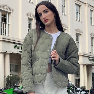 Può includere: Giacca trapuntata verde oliva con motivo geometrico. La giacca ha un colletto tondo e chiusura con bottoni. La persona indossa un top bianco e pantaloni bianchi sporco. Lo sfondo include un edificio con colonne e la scritta "WAIT & PAT".