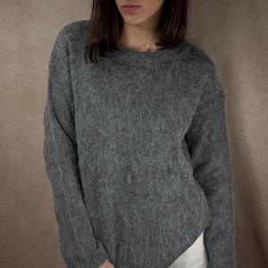 May include: A gray, long-sleeved sweater with a crew neck. The sweater has a fuzzy texture and is paired with white pants. The model is standing in front of a brown background.