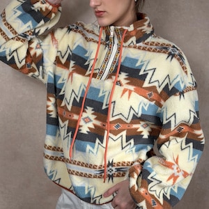 May include: Cream fleece pullover with a geometric pattern in brown, blue, and grey. It has a high collar, half-zip, and orange drawstrings. The long sleeves and relaxed fit make it a comfortable garment. The design is reminiscent of a tribal pattern.