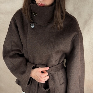 May include: A dark brown wool coat with a high, buttoned collar and a matching belt. The coat has a tailored silhouette with long sleeves and a wrap-around design. The fabric appears soft and textured. The coat's design is minimalist and elegant.