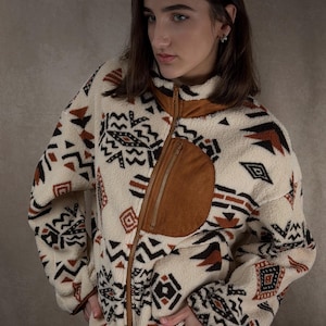 Pode incluir: Casaco de lã polar creme com um padrão geométrico castanho e um bolso de camurça castanho. O casaco tem gola alta e fecho de correr completo. O design inclui formas pretas, castanhas e creme. Roupa exterior quente e elegante.