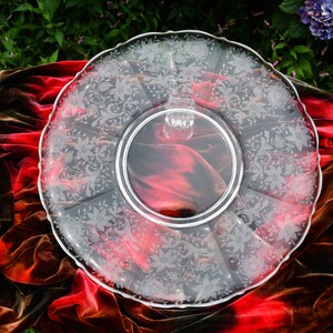 May include: A clear glass serving platter with a scalloped edge and an etched floral design. The platter is on a red velvet cloth.