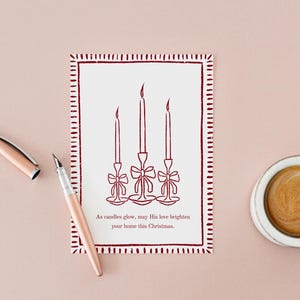 K&ouml;nnte beinhalten: Eine Weihnachtskarte mit rotem Rand und einer Zeichnung von drei Kerzen, einem Stift und einer Tasse Kaffee auf rosa Hintergrund. Auf der Karte steht: &bdquo;M&ouml;ge seine Liebe Ihr Zuhause an diesem Weihnachten erhellen.&ldquo;