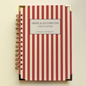 May include: A spiral-bound daily planner with a red and cream vertical striped cover. The planner features gold corner protectors and a gold spiral binding. The cover includes the text "ABIDE & ACCOMPLISH - DAILY PLANNER - worry less, worship more."