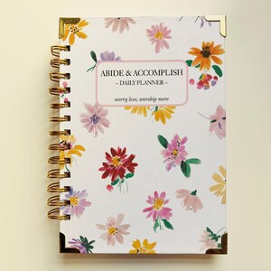 May include: A floral-patterned daily planner with a gold spiral binding and corner protectors. The white cover is decorated with watercolour flowers in pink, yellow, and red. Text on the cover reads "ABIDE & ACCOMPLISH - DAILY PLANNER - worry less, worship more."