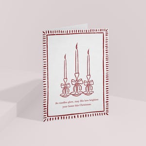 K&ouml;nnte beinhalten: Eine gefaltete Weihnachtskarte mit wei&szlig;em Hintergrund und einem weinroten Rand. Die Karte zeigt eine Strichzeichnung von drei Kerzen mit Schleifen. Der Text unten lautet: &bdquo;M&ouml;ge seine Liebe Ihr Zuhause an diesem Weihnachten erhellen.&ldquo;