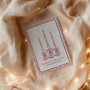 K&ouml;nnte beinhalten: Weihnachtskarte mit roter Umrandung und einer Zeichnung von drei Kerzen auf wei&szlig;em Hintergrund. Der Text lautet: &bdquo;M&ouml;ge seine Liebe Ihr Zuhause an diesem Weihnachten erhellen.&ldquo; Auf beigem Stoff mit Lichterketten.