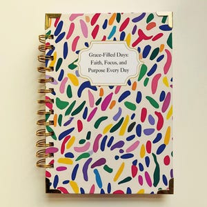 Op de afbeelding: Een spiraalgebonden notitieboek met een witte kaft versierd met kleurrijke, abstracte vormen in tinten blauw, groen, roze, geel en paars. De kaft bevat de tekst "Grace-Filled Days: Faith, Focus, and Purpose Every Day."