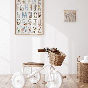 May include: White tricycle with a brown seat and a wicker basket. The tricycle is in a room with a white wall and a framed alphabet poster with illustrations.