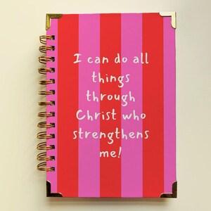 May include: A notebook with a red and pink striped cover and gold spiral binding. The cover has the text "I can do all things through Christ who strengthens me!" in white. Gold corner protectors are also visible.