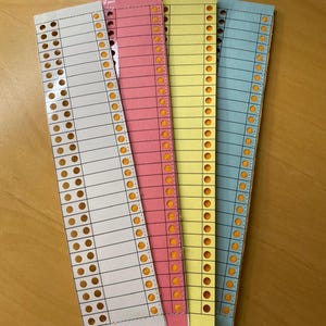 May include: Four coloured paper strips in white, pink, yellow, and blue. Each strip has a series of small holes along one edge and horizontal lines for writing. The paper is on a light brown wooden surface.