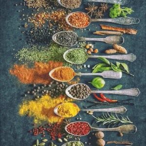 May include: A collection of various spices and herbs in silver spoons on a dark blue background. The spices include cloves, coriander seeds, red pepper flakes, black peppercorns, dried herbs, turmeric, paprika, cumin, and peppercorns.