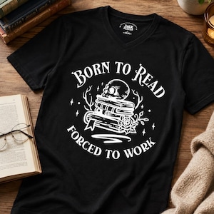 Könnte beinhalten: Ein schwarzes V-Ausschnitt-T-Shirt mit einem weißen Grafikdruck eines Bücherstapels, der von einem Totenkopf und einer Rose gekrönt wird. Der Text "BORN TO READ" ist über den Büchern und "FORCED TO WORK" darunter gebogen, ergänzt durch kleine Sterne.