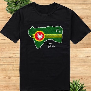 May include: Black t-shirt with a green and yellow design of the flag of the island of Tain, featuring a white chicken in a red circle. The text "Tain" is printed below the design.